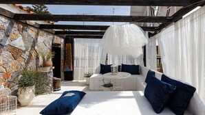 Un espace de détente extérieur accueillant, avec un mur en pierre, une table blanche, des coussins bleus et un auvent blanc suspendu.