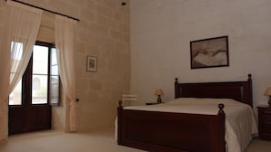 A bedroom with a wooden bed, a window with curtains, a nightstand with a lamp, and a painting on the wall. - Gozo Holiday Farmhouse Accommodation (Gharb Gozo)