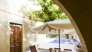 An outdoor patio with a stone floor, a wooden table with chairs, and a white umbrella providing shade. - Gozo Holiday Farmhouse Accommodation (Gharb Gozo)