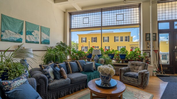 A cozy living room with a large window, a sofa set, a coffee table, and various potted plants.