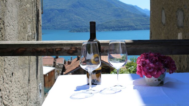 A table with a wine bottle and two glasses, a potted plant, and a scenic view of a lake and mountains. - 2 1/2-room vacation apartment in Vercana, quiet and inviting above Lake Como (Vercana)