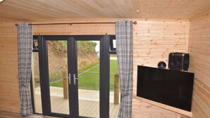A room with a large sliding glass door leading to an outdoor deck, a flat-screen TV mounted on the wall, and a speaker on a wooden shelf.