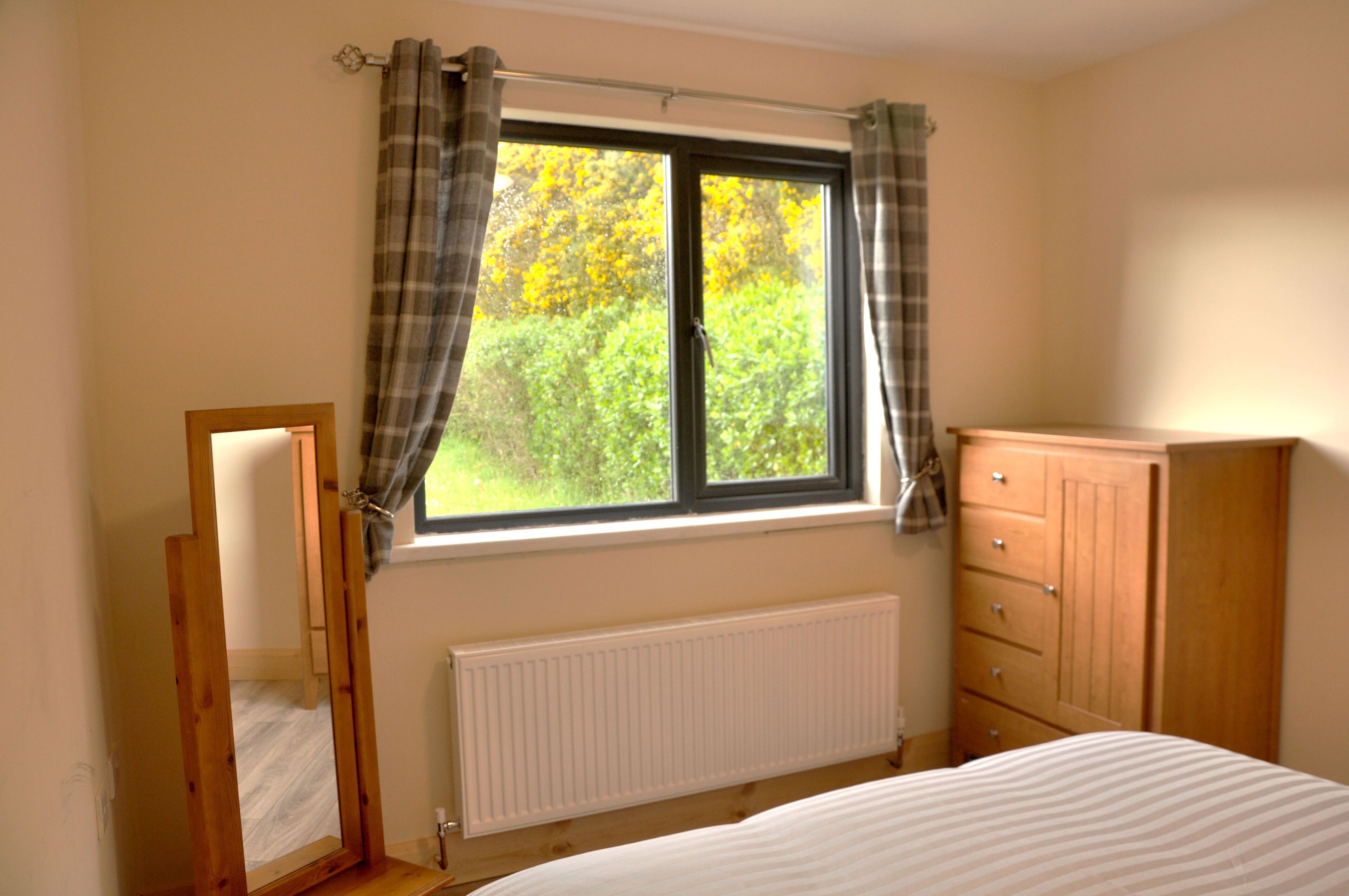 A bedroom with a window, a radiator, a wooden wardrobe, and a mirror.
