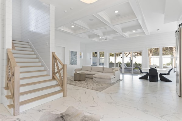 A spacious living area with a wooden staircase, a beige sectional sofa, and a large window offering a view of the outdoors.