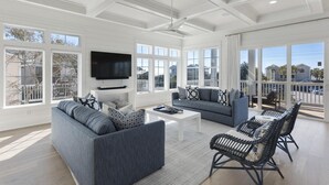 Ein geräumiges Wohnzimmer mit einem großen Fernseher, grauen Sofas, Korbsesseln und großen Fenstern mit Blick auf die Umgebung.