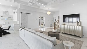 Ein geräumiges Wohnzimmer mit einem großen, grauen Sofa, einem Couchtisch und einem an der Wand montierten Fernseher. Eine Schiebetür führt zu einem Balkon, und die Decke ist weiß mit integrierter Beleuchtung.