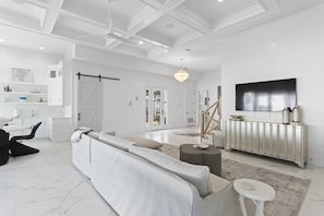 A spacious living room with a large grey sofa, a coffee table, and a TV mounted on the wall. There is a sliding door leading to a balcony, and a white ceiling with recessed lighting.