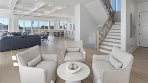 Ein geräumiges Wohnzimmer mit grauem Sofa, zwei Sesseln und einem Couchtisch. Eine Treppe mit Holzhandläufen führt zu einer oberen Ebene.
