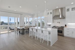 A modern kitchen with white cabinetry, a central island with bar stools, a stainless steel range hood, and a view to the outdoors.