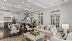 A spacious living room with a high ceiling, white walls, a central staircase, and a kitchen area with modern appliances.