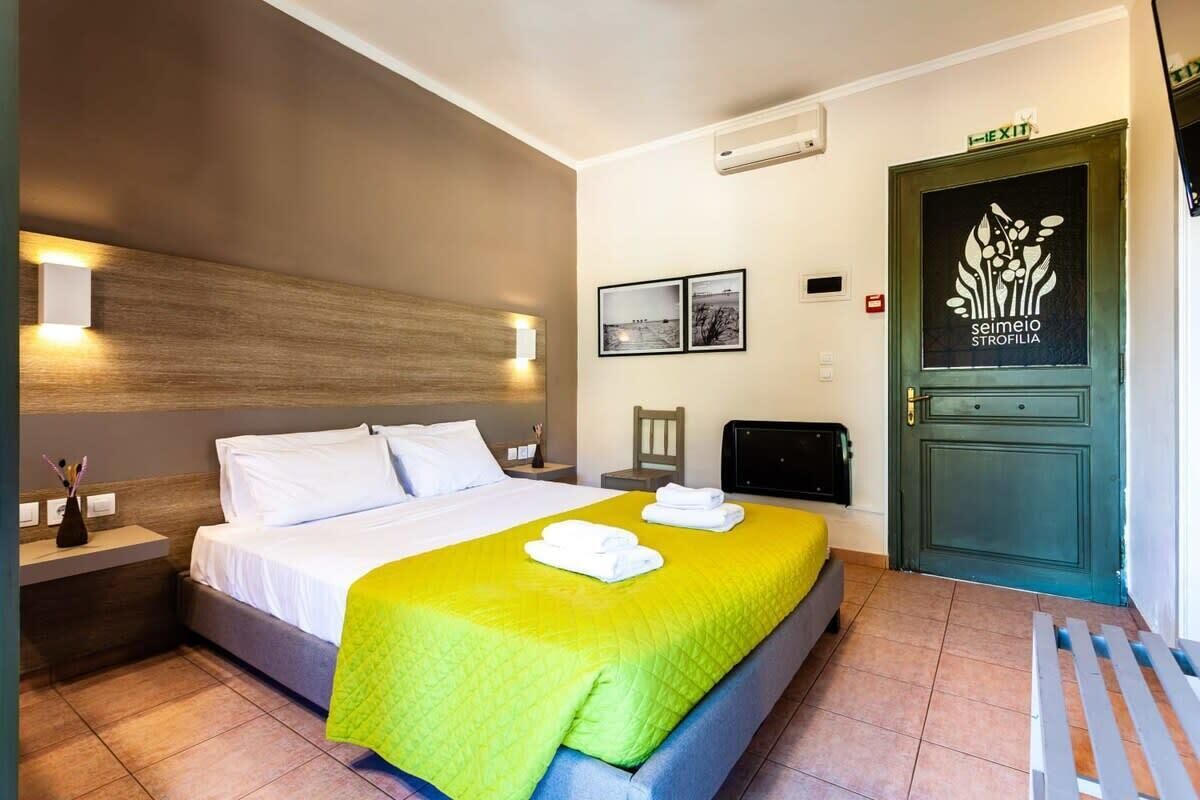 A hotel room with a bed, a nightstand, a desk, a chair, a door with a logo, and a wall-mounted air conditioner.
