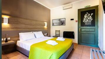 A hotel room with a bed, a nightstand, a desk, a chair, a door with a logo, and a wall-mounted air conditioner.