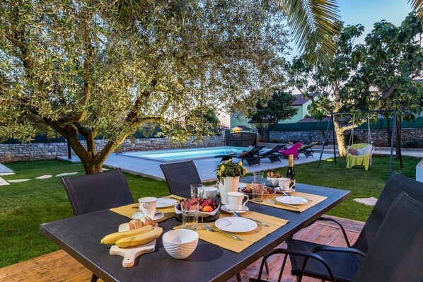 Un área de comedor al aire libre con una mesa dispuesta para una comida, rodeada de sillas y una piscina al fondo.