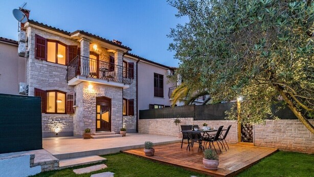 A two-story house with a stone facade, a balcony with a railing, and a wooden deck with outdoor furniture.