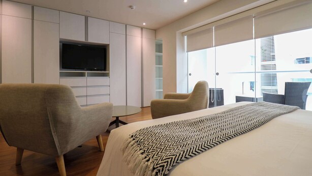 A modern hotel room with a large bed, two armchairs, a TV, and a balcony with a view of the city.