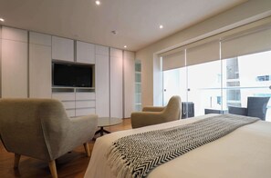 Ein modernes Hotelzimmer mit einem großen Bett, zwei Sesseln, einem Fernseher und einem Balkon mit Blick auf die Stadt.