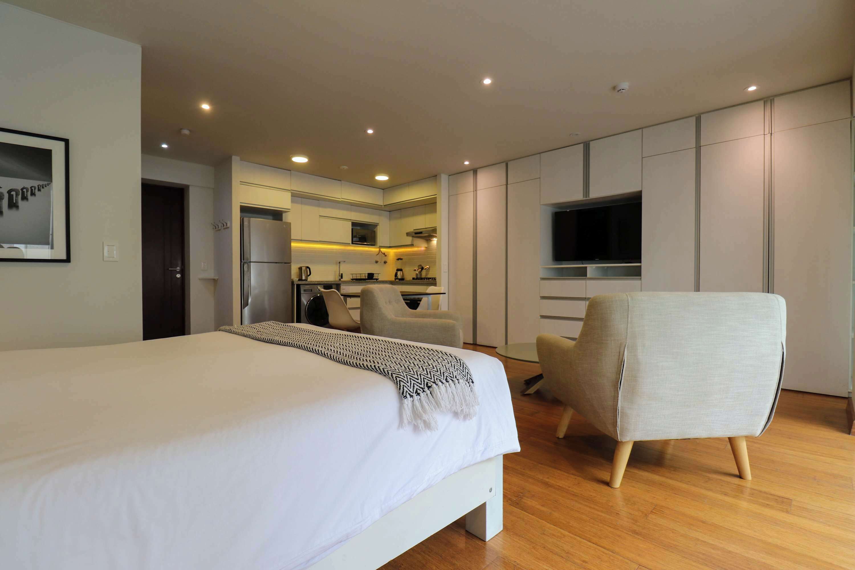 Une chambre d’hôtel moderne dotée d’un grand lit, d’un coin salon avec un fauteuil, d’une kitchenette et d’un téléviseur fixé au mur.