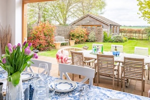 An outdoor dining area with a table set for two, surrounded by wooden chairs, a wooden table, and a view of a garden with a wooden building and trees.