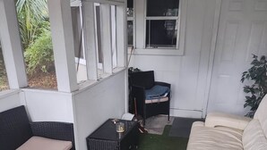 Un patio techado con césped artificial verde, un sofá blanco, una silla de mimbre negra y una mesita con un vaso y una botella.