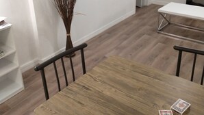 Un comedor con una mesa de madera, sillas y un jarrón con ramas. Se ve una puerta al fondo y un cuadro colgado en la pared.
