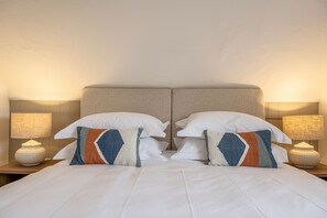Ein Doppelbett mit weißen Leinen, zwei Zierkissen und zwei Nachttischlampen auf Holz-Nachttischen.
