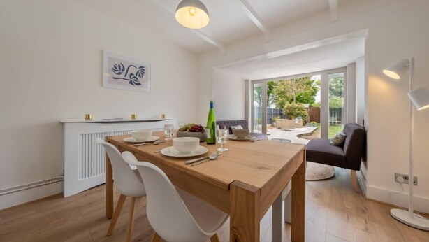 Uma área de jantar moderna com mesa de madeira, cadeiras brancas e uma luminária pendente. Há uma janela com vista para o verde lá fora.