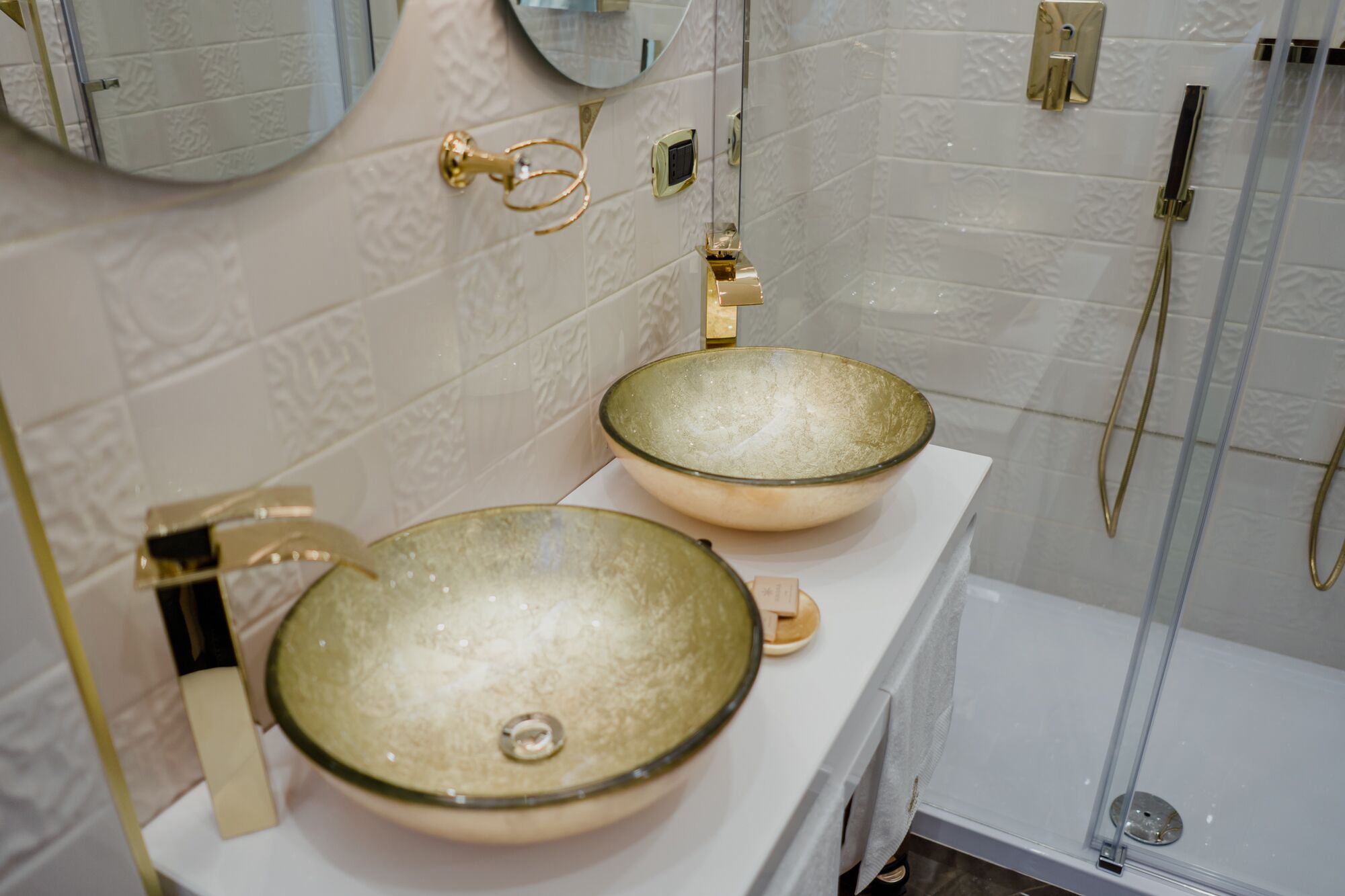 Un baño con dos lavamanos grandes y redondos de cerámica color beige con detalles dorados, un espejo encima y una zona de ducha con paredes de vidrio.