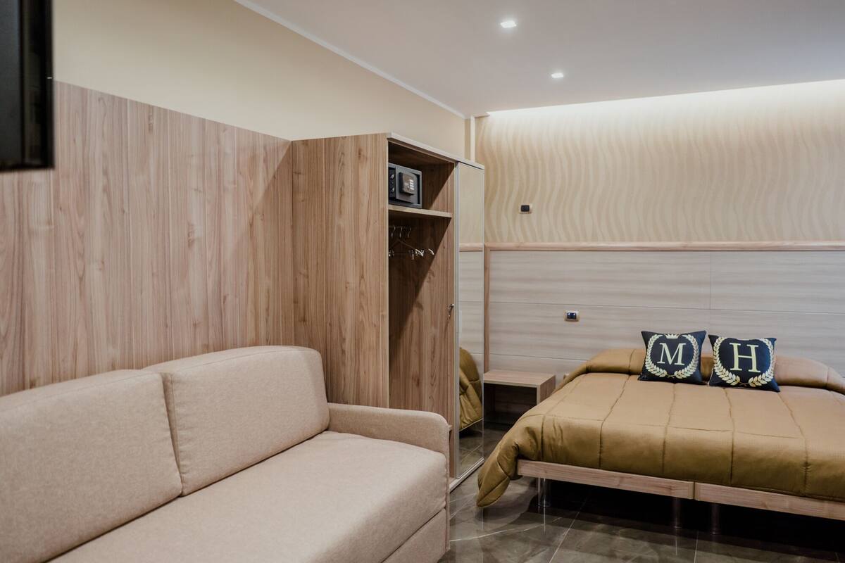A modern bedroom with a beige sofa, a wooden wardrobe, and a bed with two pillows.