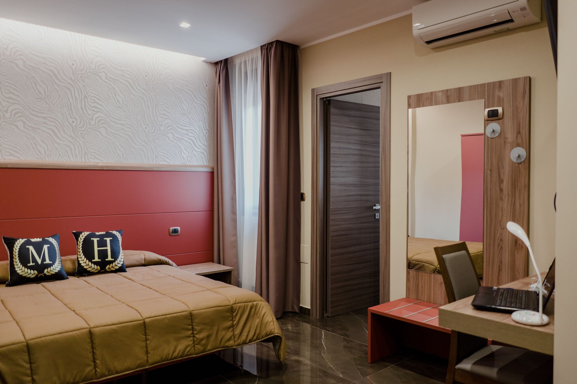 A hotel room with a bed, a desk with a laptop, an air conditioner, a door, and a mirror.