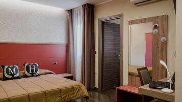 A hotel room with a bed, a desk with a laptop, an air conditioner, a door, and a mirror.
