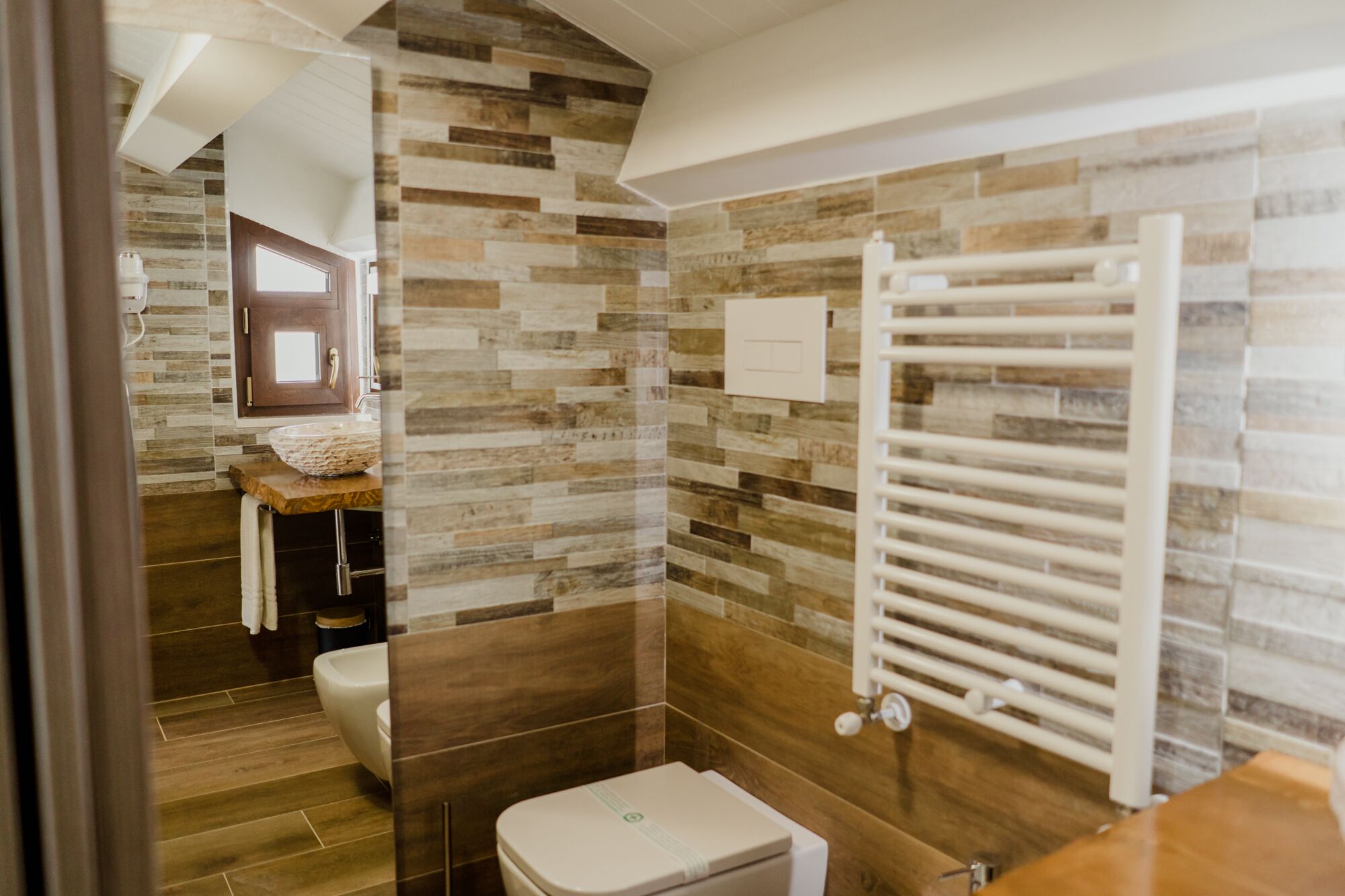 Ein modernes Badezimmer mit einer Wand aus Steinmosaik, einem Holzlager für Waschtischutensilien, einer weißen Toilette und einem weißen Handtuchhalter.