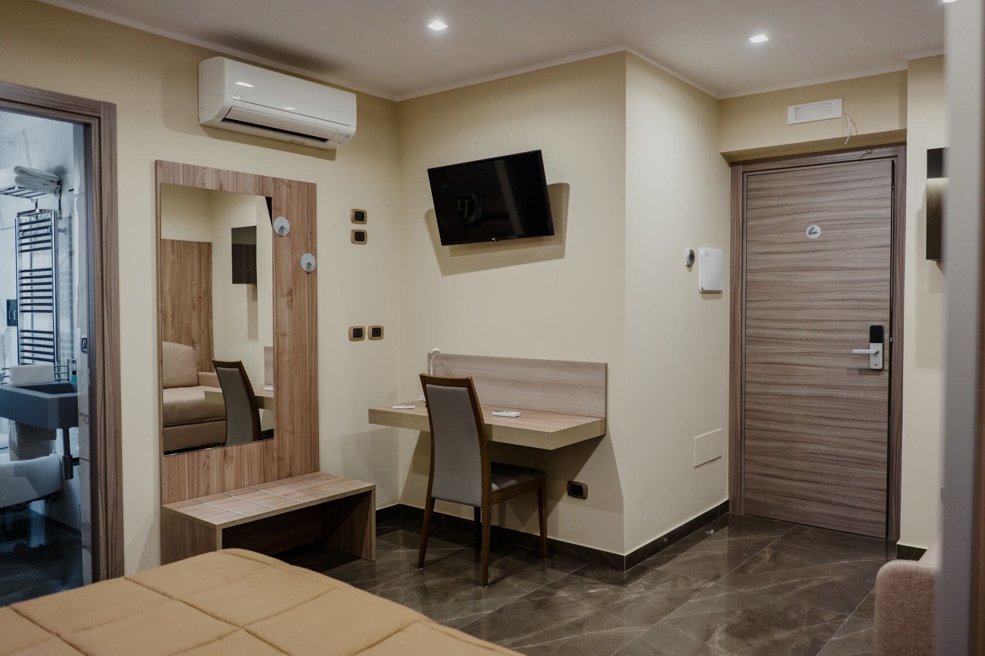 Een moderne hotelkamer met een bed, een bureau met stoel, een televisie, een airconditioningsysteem en een deur.