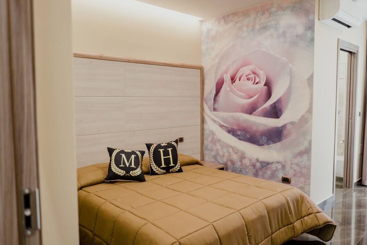 A bedroom with a bed, two pillows with 'MH' initials, a large floral mural, and a wooden headboard.