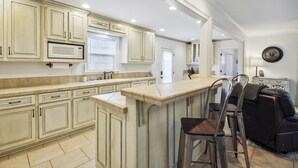 Una cocina con armarios de tonos claros, una isla central con taburetes y un comedor con sofá negro.