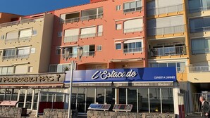 En byggnad med flera våningar, där restaurangen ”L’Estacade” ligger på bottenvåningen. Byggnaden har en fasad av sten och erbjuder uteservering.