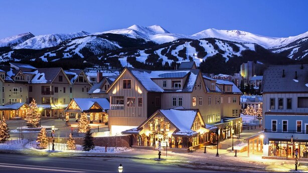 Un pueblo cubierto de nieve al anochecer, con edificios iluminados y árboles de Navidad.