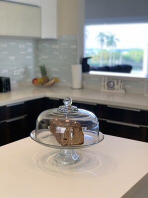 A kitchen with a clear glass cake stand on a white countertop, a microwave, and a window with a view of palm trees. - Beautiful Full House Rental in Private Gated Community,  Montego Bay, Jamaica (Montego Bay)
