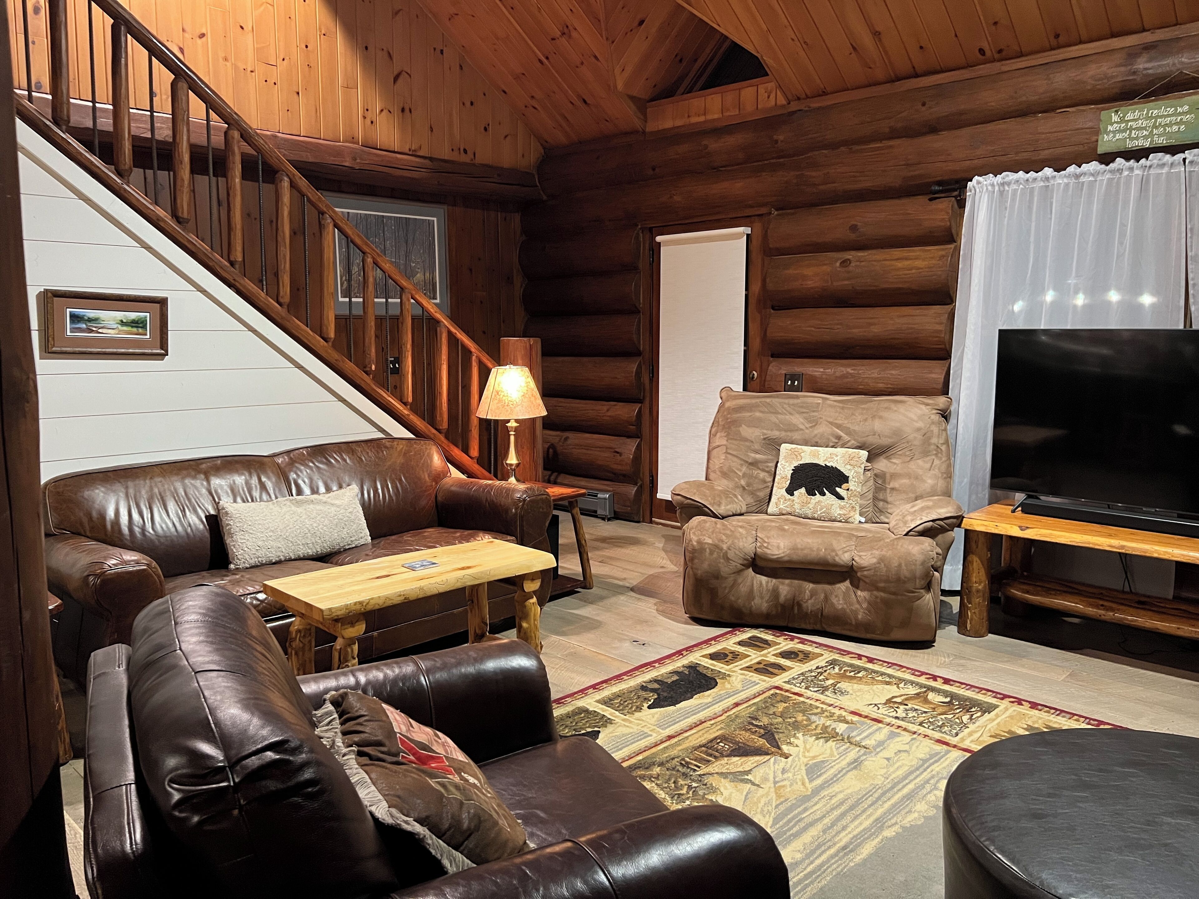 L’intérieur chaleureux d’un chalet en bois présente un canapé en cuir, une table basse en bois, un tapis à motifs, un meuble TV en bois et un escalier menant à une mezzanine.