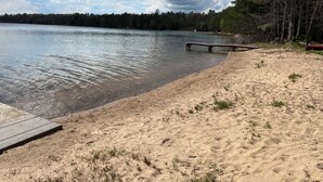 Una playa arenosa con un muelle de madera que se adentra en aguas tranquilas, rodeada de árboles frondosos.