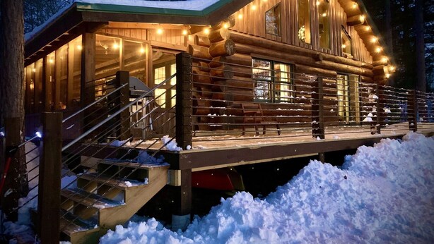 Una cabaña de troncos con una terraza techada, escaleras de madera y suelo cubierto de nieve.