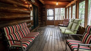Interior de una cabaña de troncos con piso de madera, asientos tapizados a rayas y grandes ventanas que ofrecen vistas del bosque circundante.