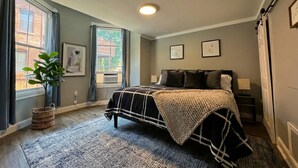 A bedroom with a bed, a window with curtains, a plant, and framed pictures on the wall. - The Grayson at Park Place - Historic Charm Meets Modern Comfort (Wilmington)