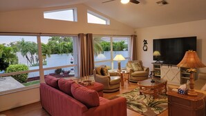 Amplia sala de estar con un sofá rojo, un sillón color beige, una mesa de centro de madera y un televisor. Amplias ventanas ofrecen vistas a un cuerpo de agua y zonas verdes.