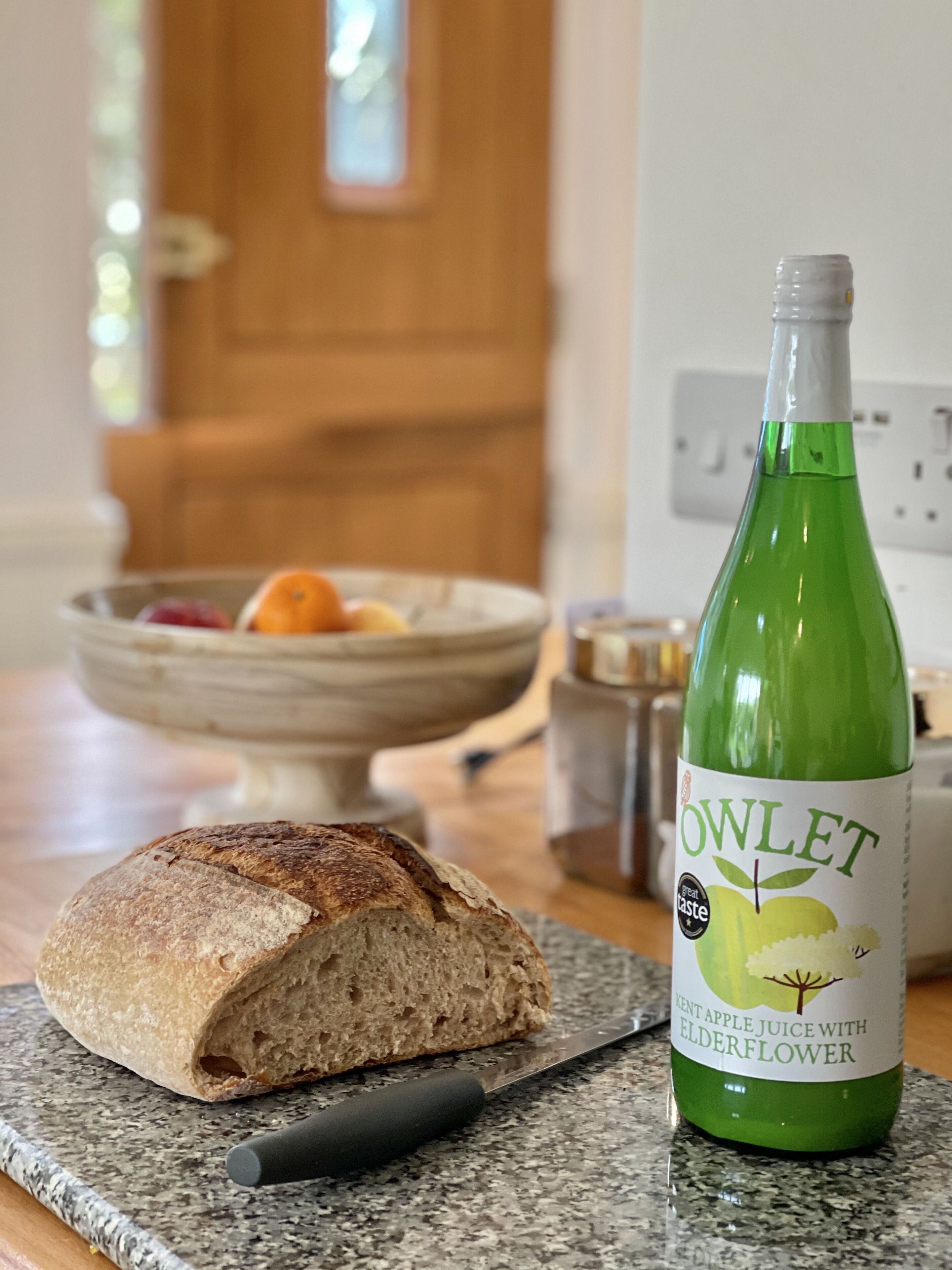 A loaf of bread on a cutting board, a knife, and a bottle of apple juice with elderflower on a kitchen counter.