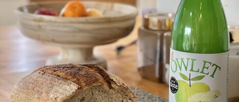 A loaf of bread on a cutting board, a knife, and a bottle of apple juice with elderflower on a kitchen counter.
