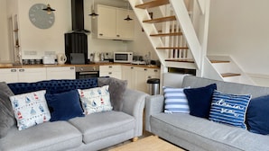 A modern living room with two grey sofas, a wooden coffee table, and a kitchen in the background. - Idyllic coastal cottage with private beach hut - Sea Rose Cottage (Kingsdown)