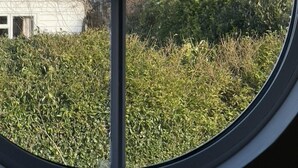 A view through a circular window of a white house with a visible front porch and a hedge in the foreground. - Idyllic coastal cottage with private beach hut - Sea Rose Cottage (Kingsdown)
