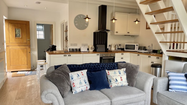 A modern kitchen with white cabinets, a built-in oven, and a microwave. There is a grey sofa with patterned cushions in the living area. - Idyllic coastal cottage with private beach hut - Sea Rose Cottage (Kingsdown)