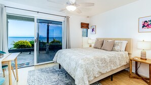 A bedroom with a large bed, a ceiling fan, a nightstand, a lamp, and a view of the ocean through a sliding glass door. - Oceanfront in Hilo Hale Kai Suites Heritage Room - Queen Bed, Private Bath (Hilo)