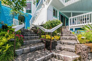 Steps lead to a modern building with a welcoming entrance, flanked by lush plants and a covered balcony area.